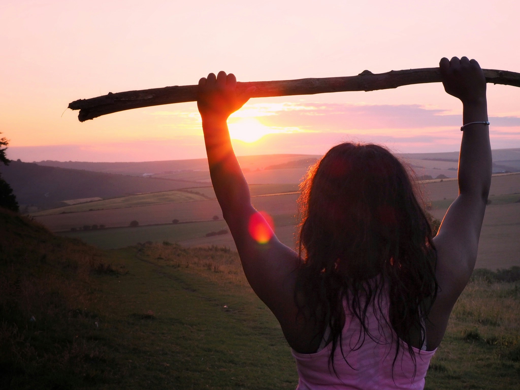 femme en haut d'une colline, vision large sur l'horizon,  dos tourné, les bras levés tenant un bâton, posture de victoire et détermination, prête à passer à l’étape suivante et à s’ouvrir au champ des possibles, symbole d’audace et de transformation : oser investir sur soi et développer sa puissance intérieure.
